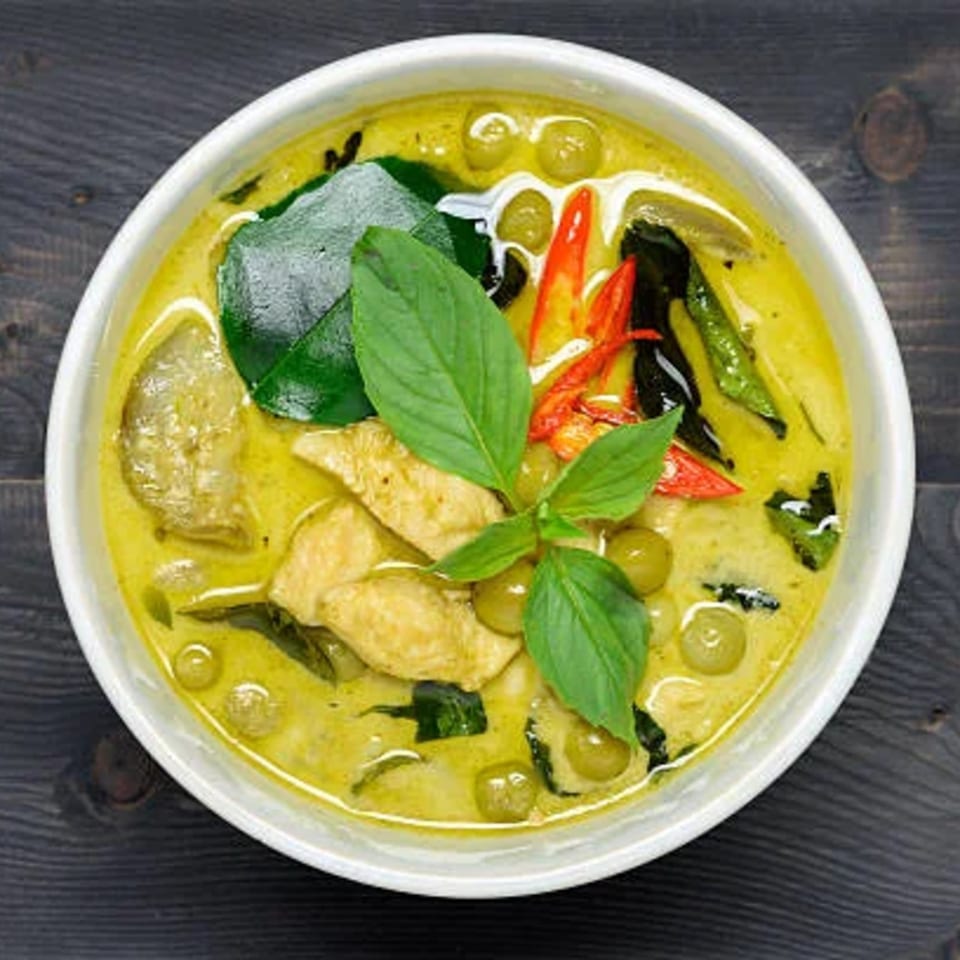 A bowl of vibrant green curry with chicken, green peas, basil, and red chili slices.